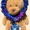 Teddy bear with a blue-purple flower lei and a small plush character hanging from it