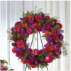 Circular floral wreath of pink roses, magenta carnations, and purple accents on a stand