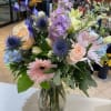 Pastel mixed bouquet with roses, gerbera daisies, and blue accents arranged in a clear glass vase