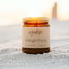 Amber jar candle labeled Midnight Poppy sitting on sand at sunset