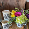 White 'I freaking love you' mugs, one filled with pink, yellow, and green flowers on a woven mat.