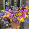 Colorful bouquet of roses, ranunculus, and purple blooms in a tan vase
