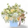 Low arrangement of white and pale yellow roses, daisies, and mixed blooms in a light blue cube vase with ribbon.