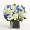 White lilies, roses, and blue flowers arranged in a square glass vase