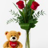 Three red roses in a glass vase beside a teddy bear holding a heart