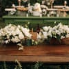 Two white floral centerpieces on a wooden table with wine glasses
