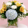 Bouquet of yellow and white flowers with a blue hydrangea in a clear glass vase