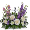White roses and purple flowers arranged around a clear glass cross.
