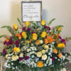 Large sympathy arrangement with yellow, orange, and purple flowers beneath a memorial sign