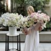 Woman holding two large pastel flower bouquets beside a white floral arrangement.