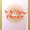 Folded pastel card with coral text 'to brighten your day' over a golden sunburst and small star accents.