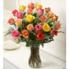 Tall glass vase filled with assorted pink, yellow, orange, and red roses with white filler flowers.