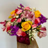 Bright mixed bouquet with yellow roses, pink carnations, and purple blooms in a pink cube vase