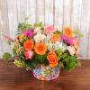 Low arrangement of orange and pink roses and gerbera daisies with blue hydrangea in a colorful round container