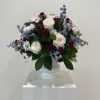 Arrangement of white roses, white daisies, burgundy carnations, and blue delphinium in a white pedestal vase