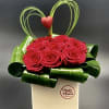 Red roses in a white box with green ribbon loops and a heart topper