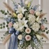 Standing floral spray with white lilies, pale roses, and blue flowers on a ribboned easel