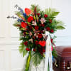 Tall funeral spray with red roses, lilies, and tropical leaves beside a casket