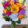 Tall mixed bouquet with sunflowers, roses, lilies, hydrangea, and purple stock in a clear glass vase.