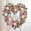 Heart-shaped standing wreath of pink roses, carnations, and white mums on a metal easel with a pink ribbon bow