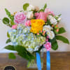 Mixed bouquet of pink and yellow roses, hydrangea, and carnations in a clear glass vase with blue ribbon