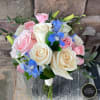 Hand-tied bouquet of ivory and pink roses with blue delphinium, wrapped in white ribbon against a stone background.