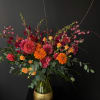 Tall arrangement of orange marigolds, pink dahlias, and spray roses in a glass vase with a gold base.