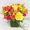 Bright arrangement of yellow roses, orange alstroemeria, and pink blooms in a green cube vase.