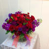 Low round arrangement of red roses and carnations with purple blooms in a clear glass vase with red and purple ribbons.