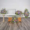 Set of pastel funeral flower arrangements surrounding an open casket in a chapel.