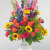 Colorful mixed bouquet in a clear glass vase with roses, sunflowers, and tall blue blooms
