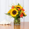Yellow sunflowers and orange roses in a clear glass vase on a table with a white seashell.