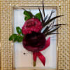 Small floral boutonniere with dark red blooms and a pink ribbon in a gold frame