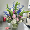 Tall mixed bouquet in a clear glass vase with pink, white, blue, and green flowers.