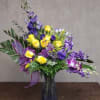 Yellow roses and purple flowers arranged in a glass vase with a ribbon