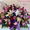 Large mixed arrangement of white lilies, pink roses, purple blooms, and red carnations with a dark purple ribbon
