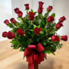 Tall arrangement of long-stem red roses with lush foliage in a dark vase with a red bow
