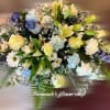Rounded arrangement of yellow roses, white lilies, daisies and blue flowers in soft pastel tones