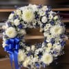 White and blue floral wreath with roses, daisies, and chrysanthemums, finished with a large royal blue bow.
