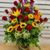 Tall mixed arrangement of red roses, sunflowers, lilies, and purple accents in a clear glass vase