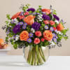 Mixed bouquet of orange roses, purple carnations, and small daisies in a round clear glass vase
