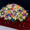 Large pastel casket spray of roses and mixed flowers in peach, purple, white, and blue on a wood casket