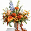 Large mixed floral arrangement in a beige vase with orange, blue, and cream blooms