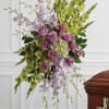 Standing spray of green gladiolus, lavender roses, and white orchids on a white easel beside a casket.