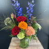 Mixed bouquet with red roses, blue delphinium, and orange blooms in a green glass vase