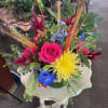 Bright mixed arrangement with hot pink rose, yellow mum, blue delphinium and wheat stems in a clear glass vase.