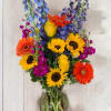 Bright mixed bouquet with sunflowers, gerberas, roses, and blue delphinium in a glass vase