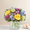 Bouquet of yellow roses, blue flowers, and pink blooms in a clear square vase