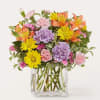 Mixed bouquet of pink, yellow, orange, and lavender flowers in a clear glass vase