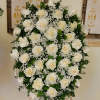 Oval standing spray of white roses and baby's breath on an easel with a black dedication sign.